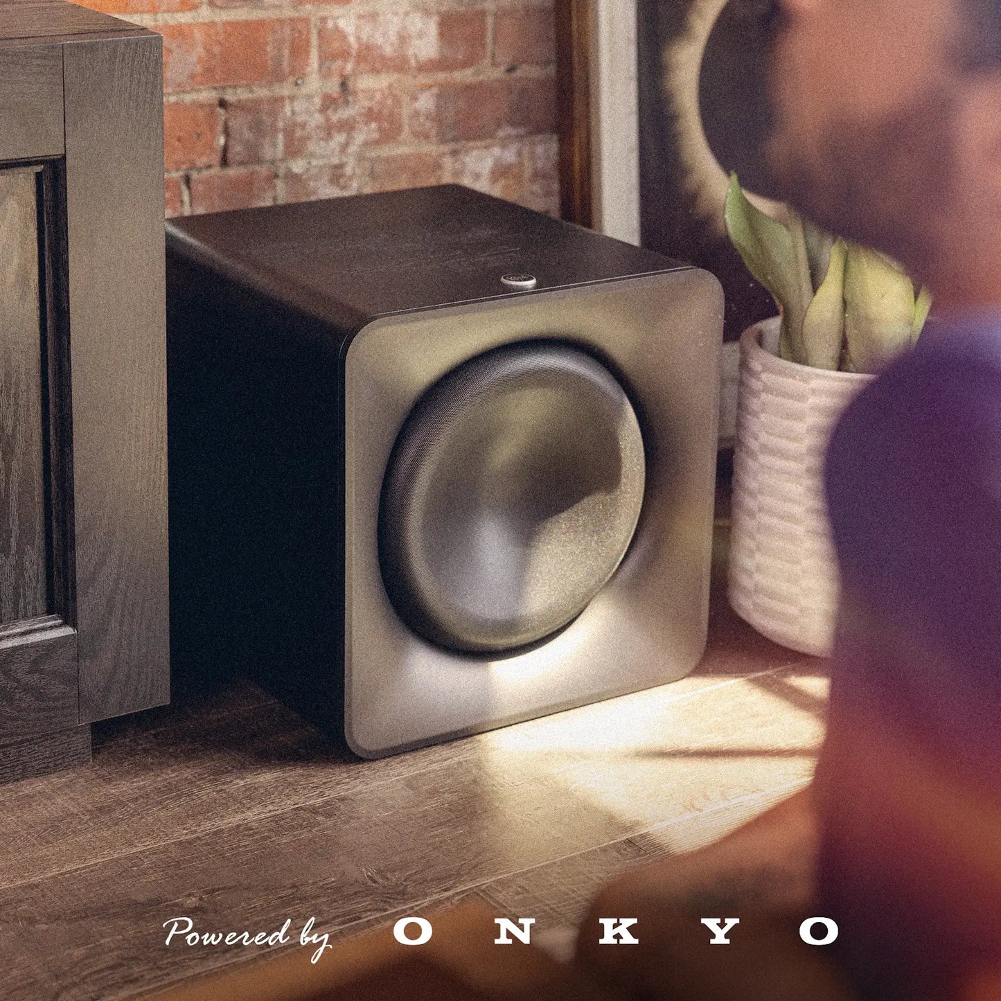 Klipsch Flexus Sub 200 aktivni nizkotonec postavljen ob sedežno garnituro v dnevni sobi za globok in močan bas. Povered by Onkyo.
